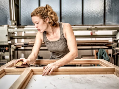 Hier sieht man Schreinermeisterin Maxime Krämer dabei, wie sie einen Holzrahmen in ihrer Werkstatt bearbeitet.